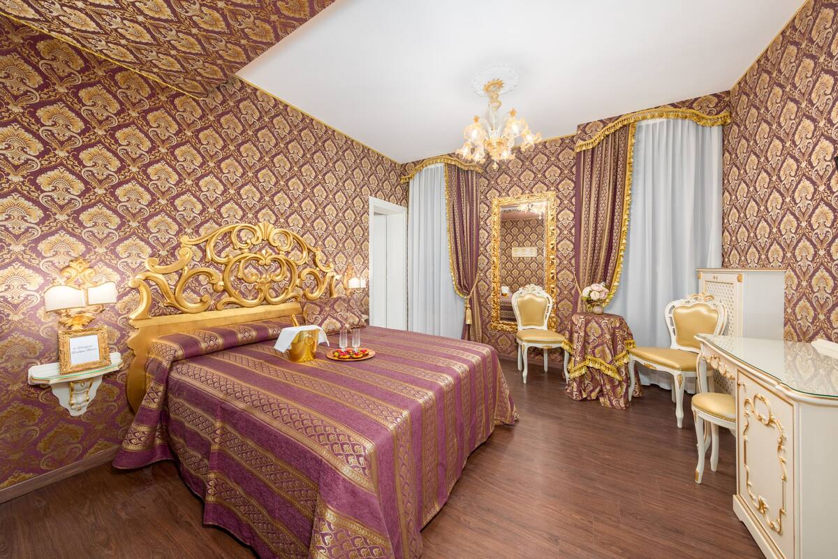 Un dormitorio lujoso con una cama grande, un cabecero ornamentado y una araña. Hay un tocador con espejo y una silla con una mesita decorativa.