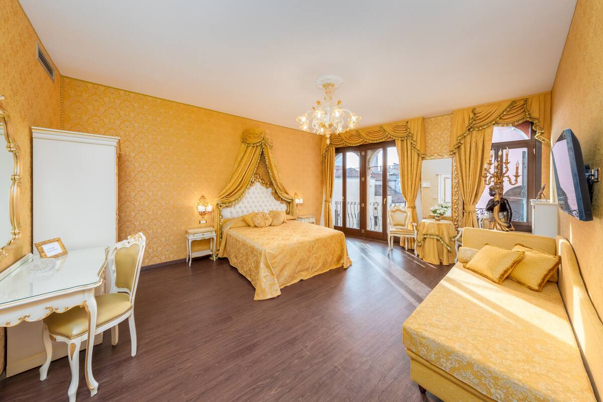 Una habitación de hotel con una cama grande, un área de descanso con un sofá, una mesa de vidrio con una silla y un televisor en la pared.
