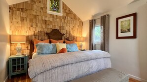 A bedroom with a wooden wall, a bed with a grey bedspread, a bench, and a nightstand with a lamp.