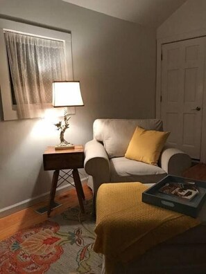 Uma sala de estar com um sofá, uma mesa lateral com abajur, um tapete pequeno e uma porta ao fundo.