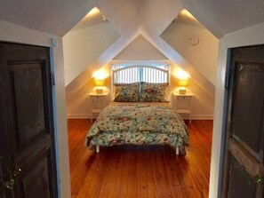 Quarto com teto inclinado, cama com roupa de cama florida, duas luminárias de cabeceira e uma janela com persianas.