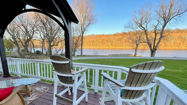 Eine Holzterrasse mit Adirondack-Stühlen und einem Glastisch, mit Blick auf einen See und Bäume.