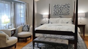 Una cama con dosel de estructura oscura, un banco a juego y una alfombra estampada. Hay dos mesitas de noche con l ámparas, una silla y un adorno en la pared.