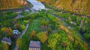 An aerial view of a cemetery surrounded by lush greenery and hills, with a bridge crossing a river in the background.