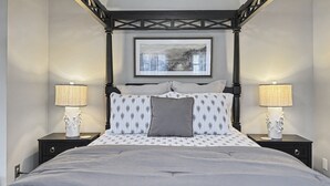 Un lit à baldaquin avec une couvre-lit grise et des coussins à motifs, flanqué de deux tables de chevet avec des lampes et une image encadrée au-dessus.