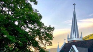 Une église dotée d’un clocher et de fenêtres en arc, entourée d’arbres et d’un mur de pierre.