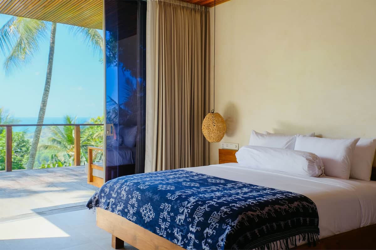 A bedroom with a large bed, a view of palm trees and the ocean, and a balcony with wooden railings.