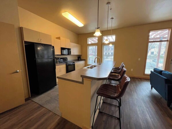 A modern kitchen with a refrigerator, stove, and sink, a bar area with stools, and large windows. - Studio Apartment at Cannery Square (Sun Prairie)
