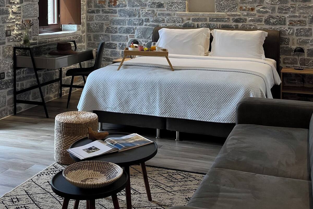 Ein gemütliches Schlafzimmer mit Steinwand, einem Bett mit weißer Bettwäsche, einem hölzernen Nachttisch mit Frühstücksangebot und einem Korbstuhl sowie einem dunkelgrauen Sofa.