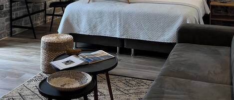 A cozy bedroom with a stone wall, a bed with white linens, a wooden bedside table with a tray of breakfast items, a wicker chair, and a dark-colored sofa.