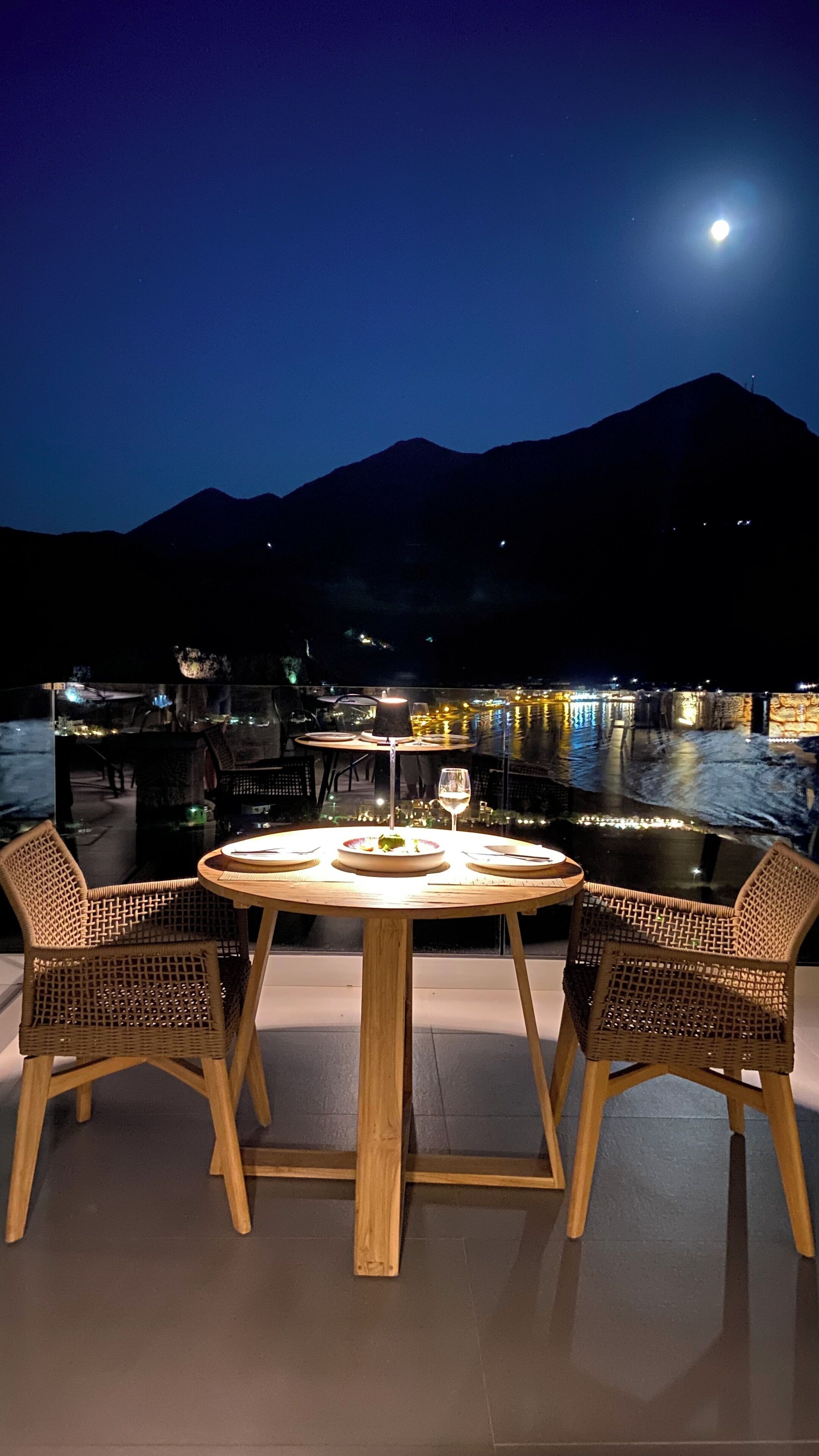A round wooden table with two wicker chairs, set outdoors at night with a view of a mountain and a lit waterfront in the background.