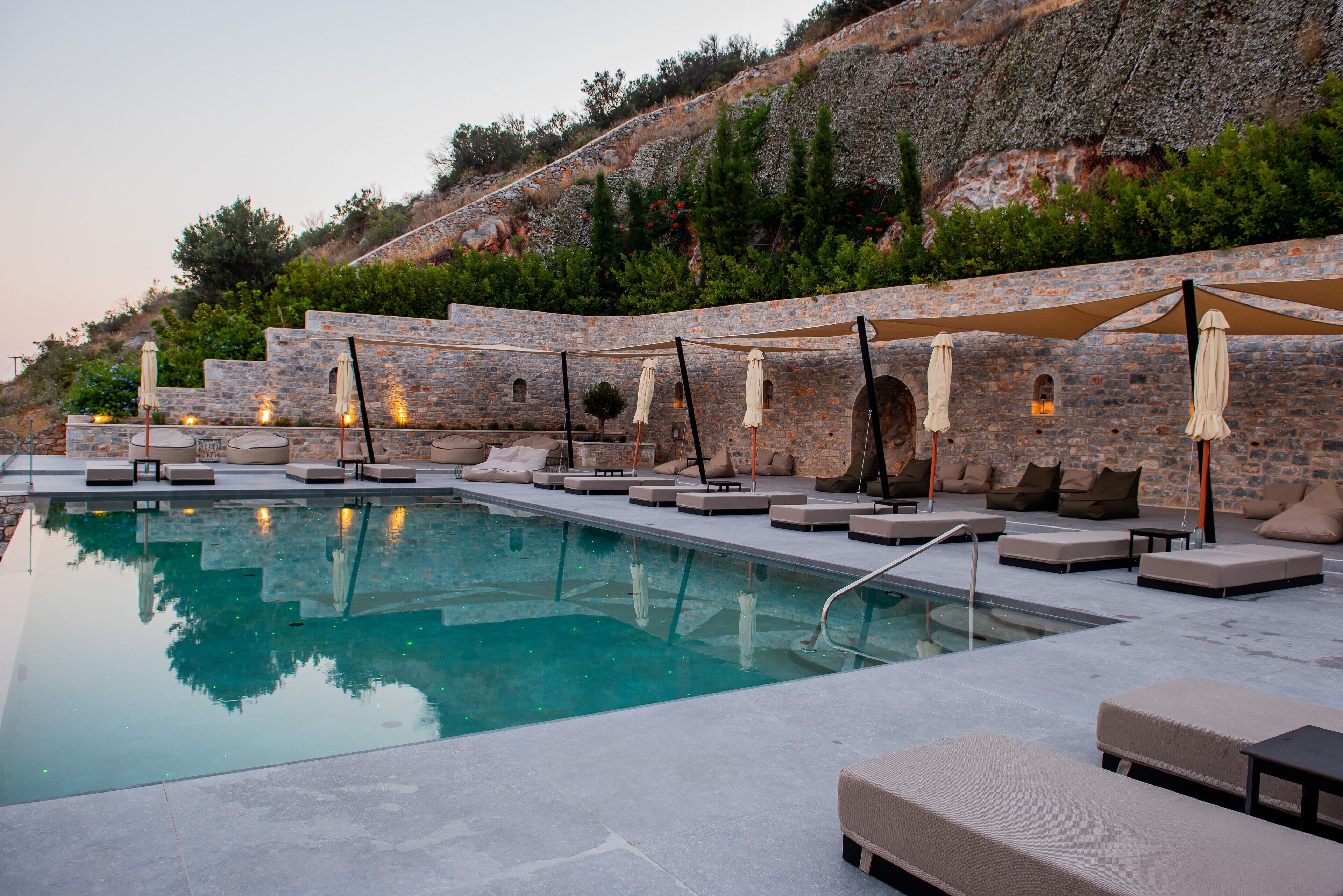 A poolside area with lounge chairs and umbrellas, set against a stone wall with a view of a hillside.