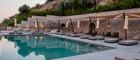 A poolside area with lounge chairs and umbrellas, set against a stone wall with a view of a hillside.