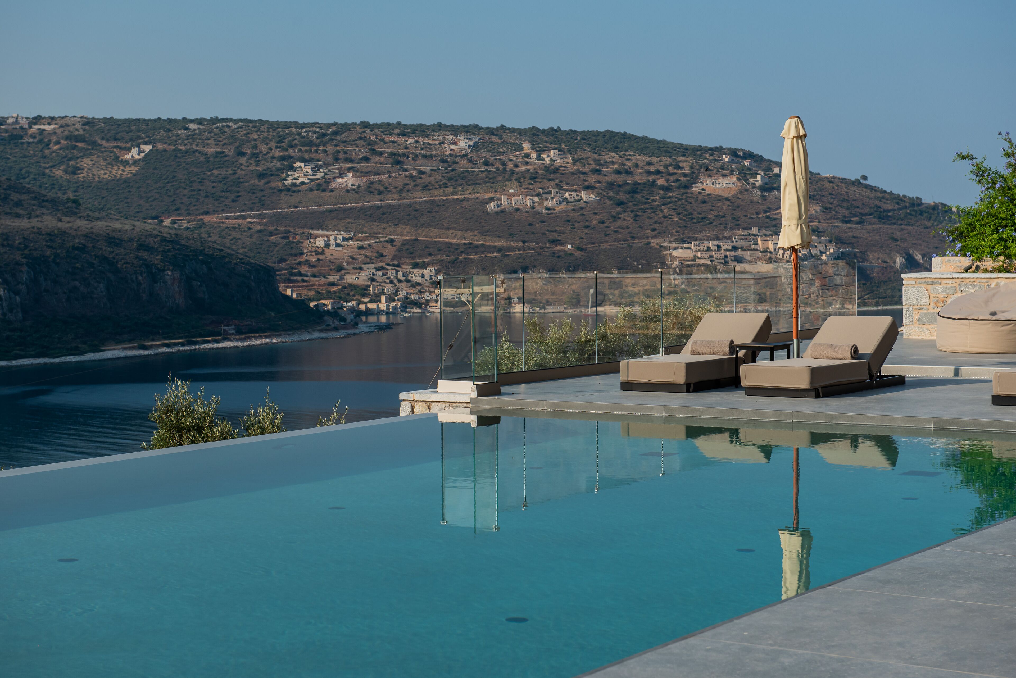 Una piscina infinita con sillones de descanso y una sombrilla, con vistas a un pueblo costero y colinas.
