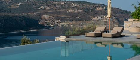 Una piscina infinita con sillones de descanso y una sombrilla, con vistas a un pueblo costero y colinas.