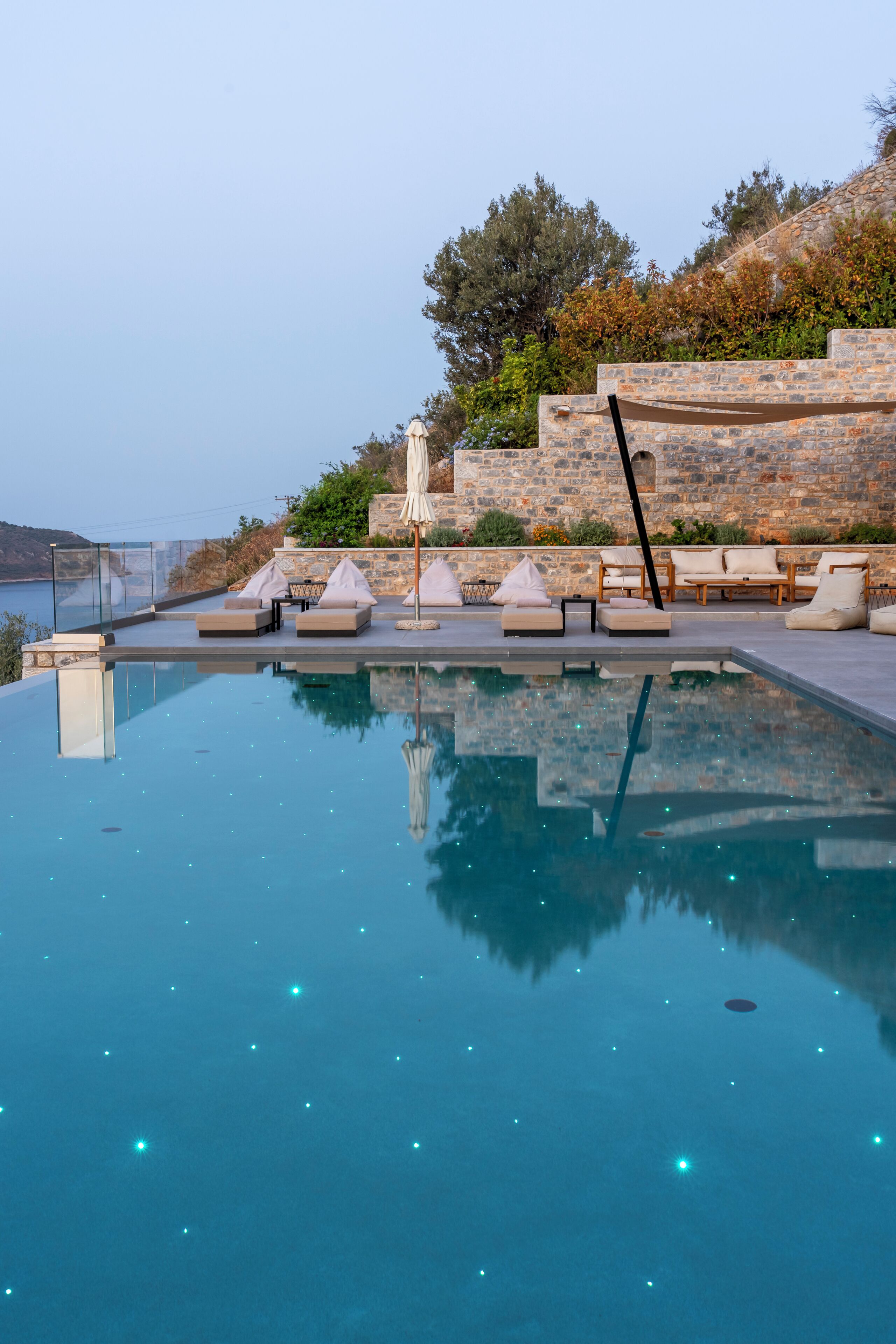 A clear blue infinity pool with lounge chairs and umbrellas, surrounded by stone walls and greenery.