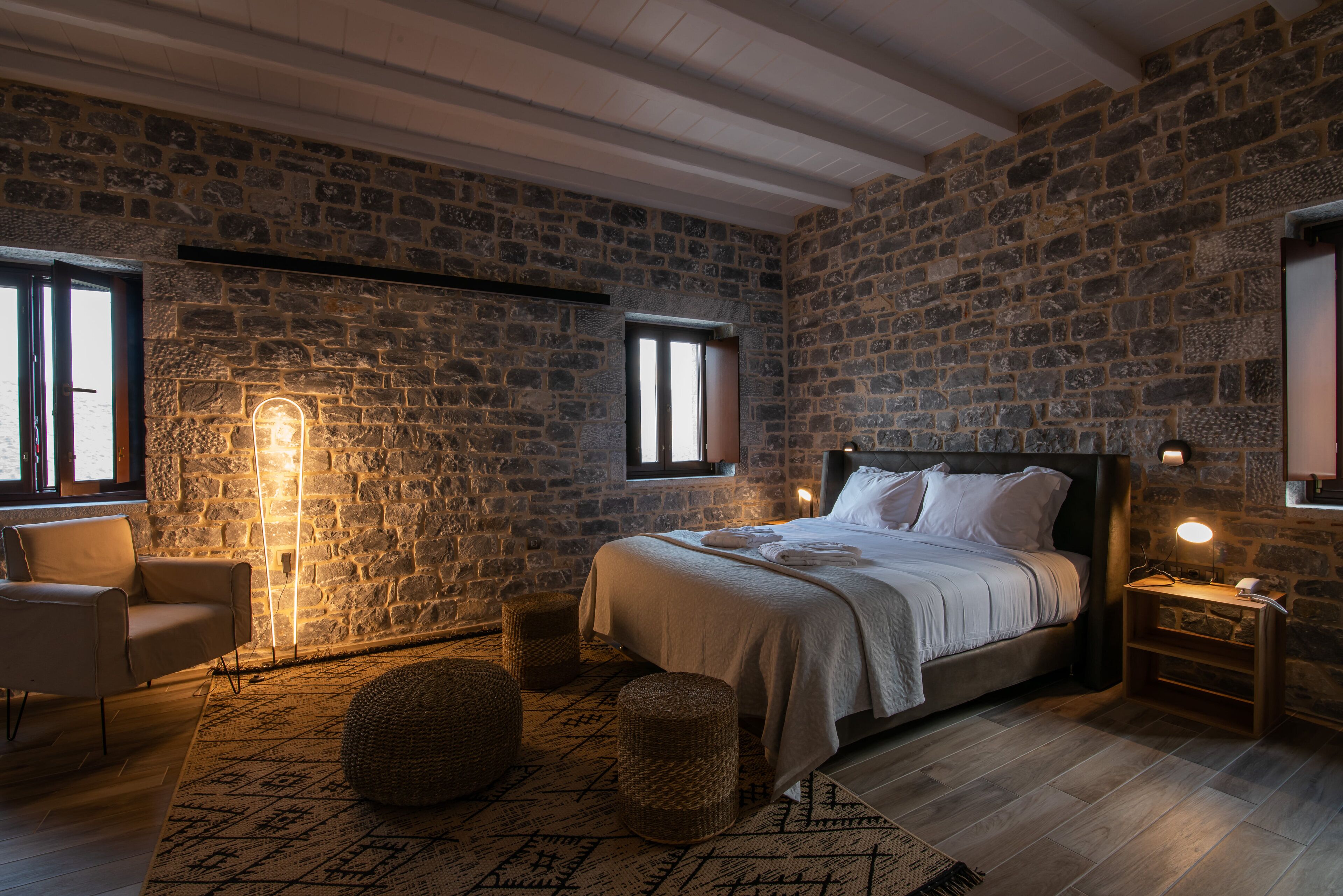 A bedroom with a stone wall, a bed with white linens, a chair, a bedside table with a lamp, and a large window.