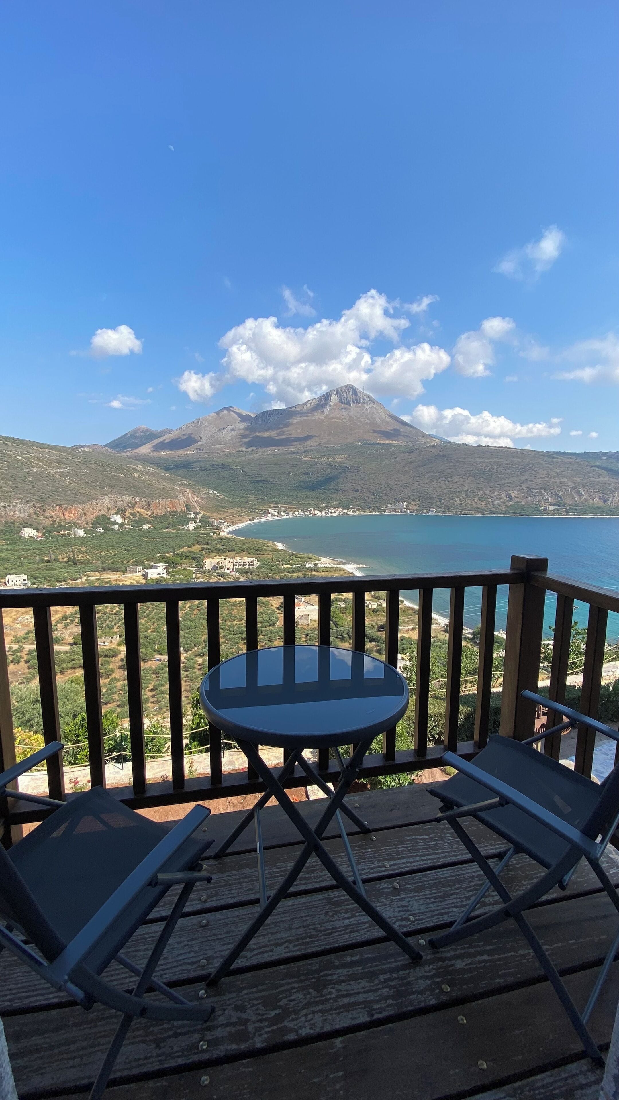 A balcony with a table and two chairs, overlooking a coastal landscape with a mountain and a beach.