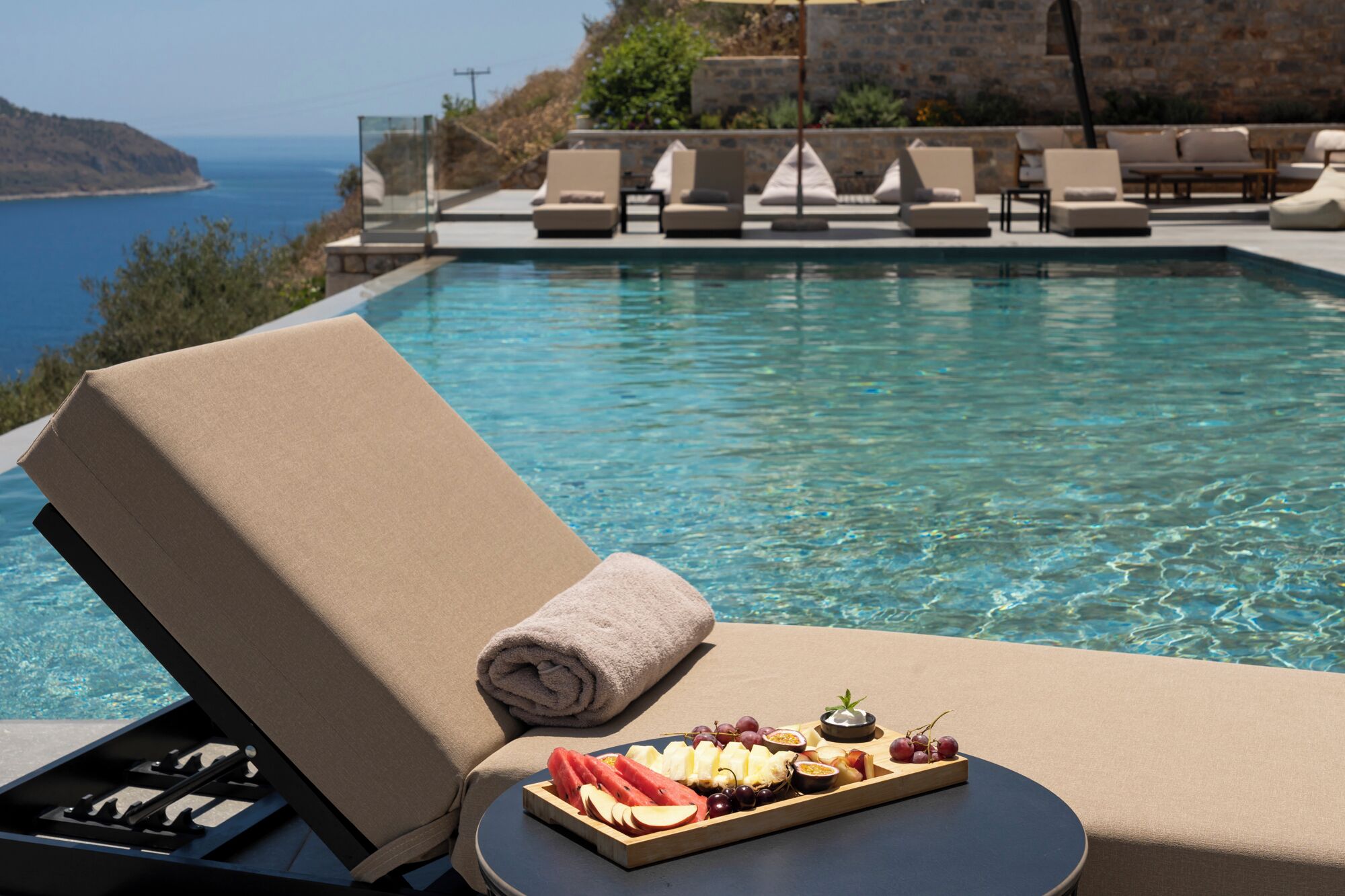 A poolside lounge with a sun lounger, a tray of fruit and cheese, and a view of the sea.