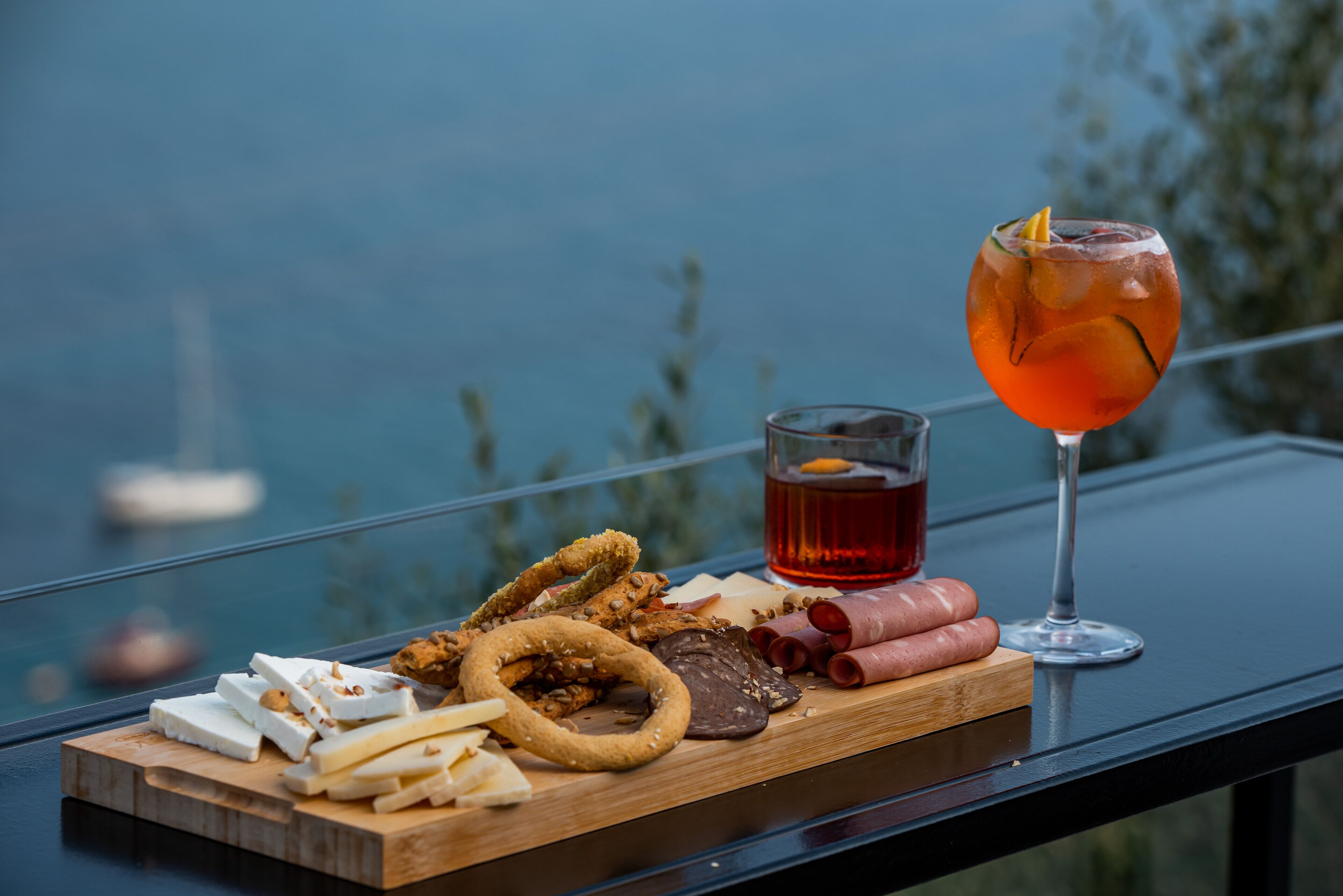 A wooden board with various cheeses, meats, and snacks on a balcony with a view of the sea.