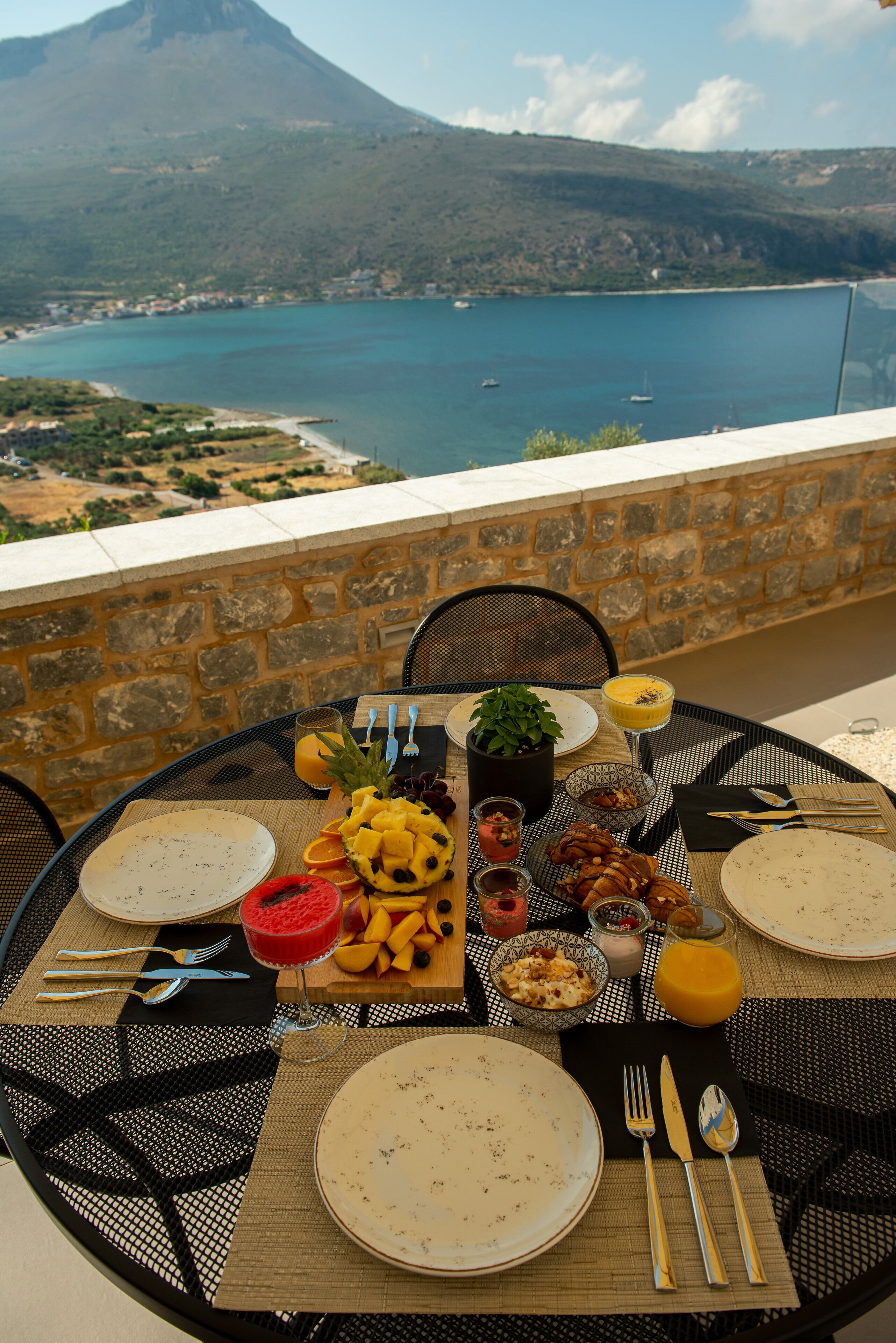 Ein Tisch ist für eine Mahlzeit gedeckt, mit Blick auf eine Küstenlandschaft und einem Berg im Hintergrund.