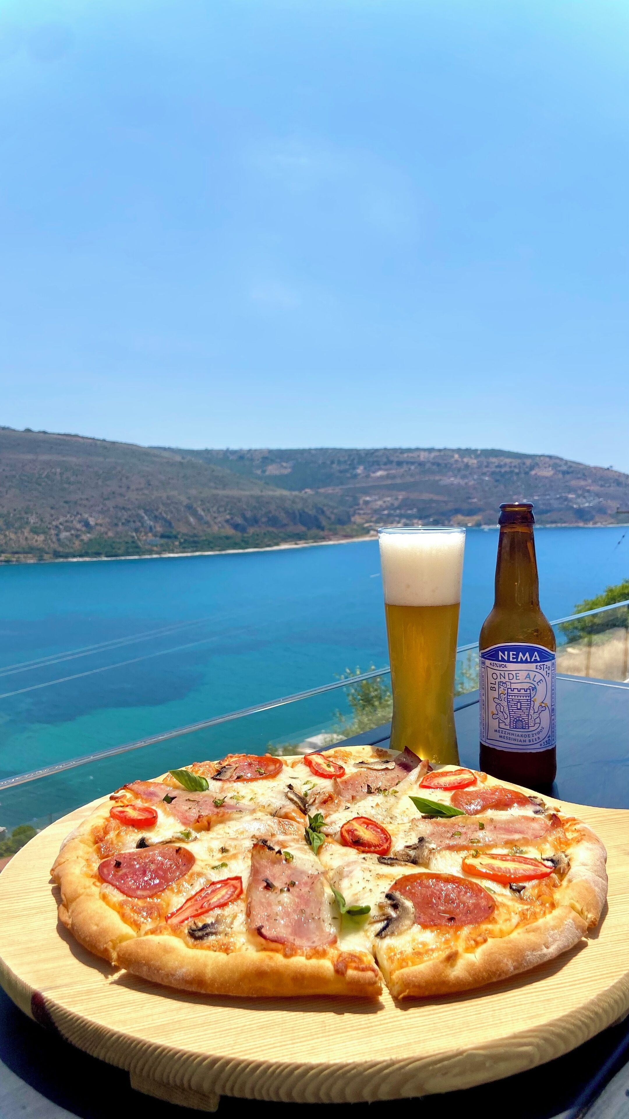 A pizza with toppings, a glass of beer, and a bottle on a table with a view of a lake and hills.