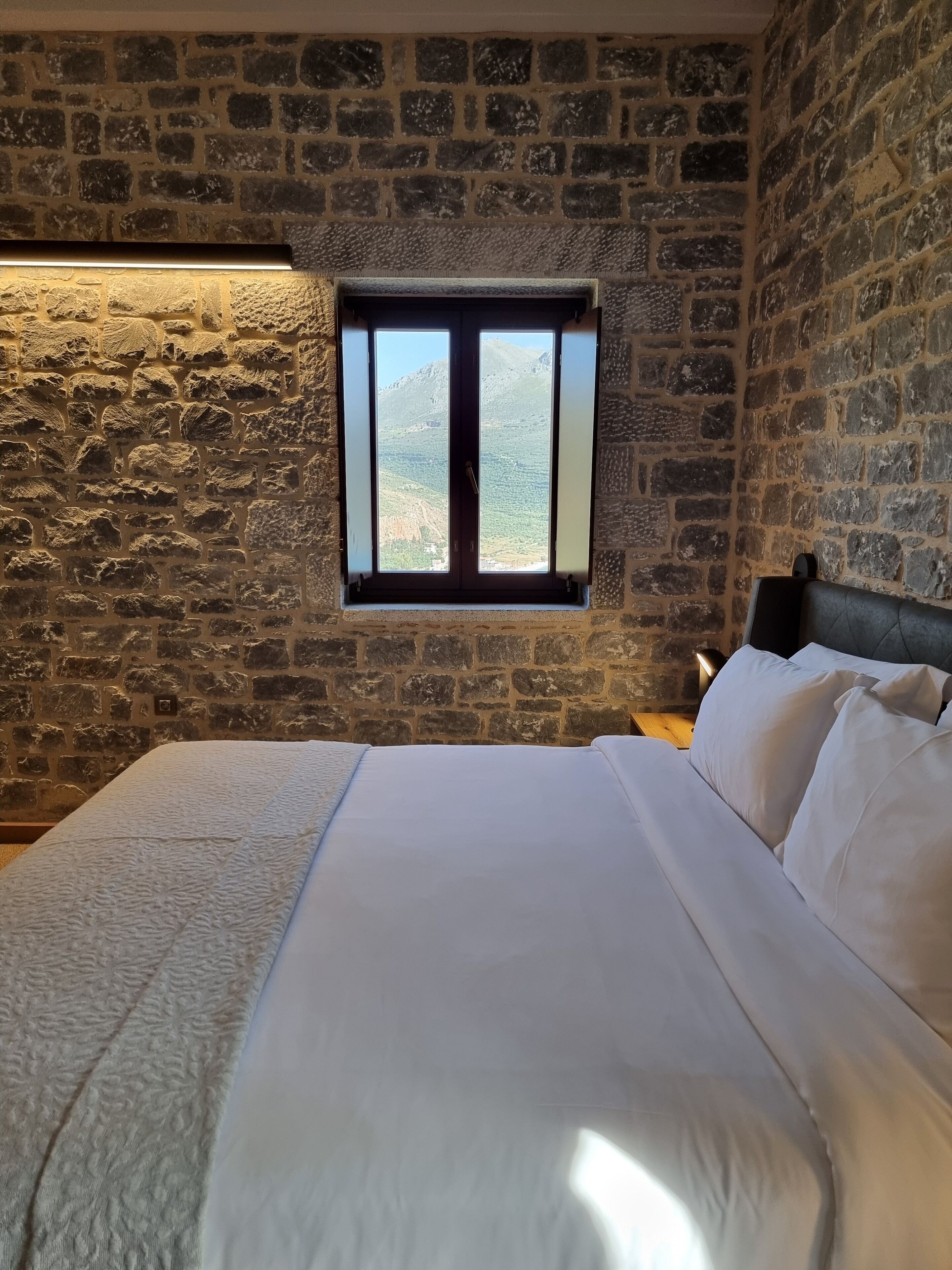 Une chambre avec un mur en pierre, une fenêtre donnant sur une montagne et un lit recouvert de draps blancs.