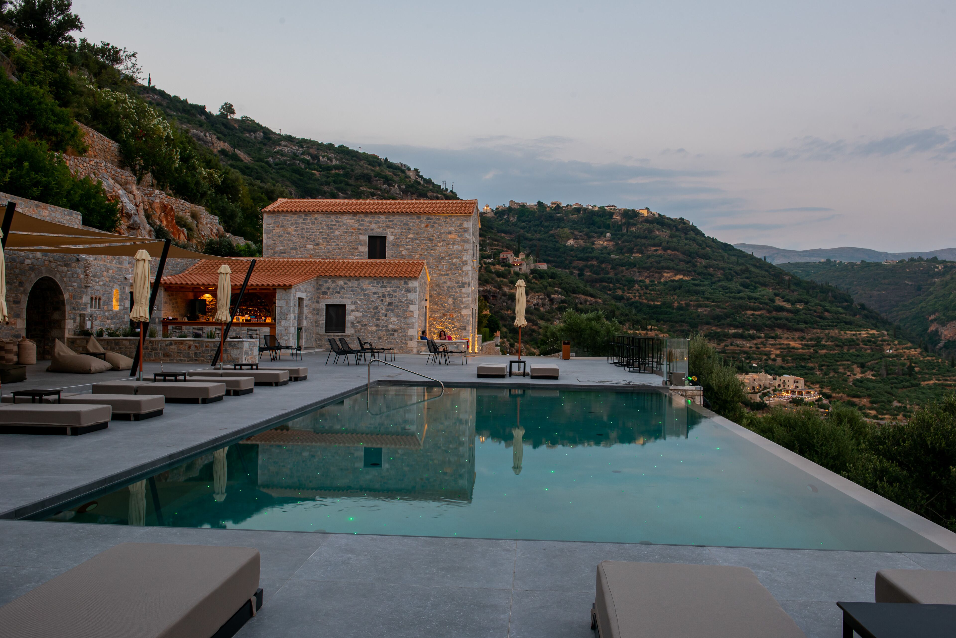 An infinity pool with lounge chairs and a stone building with a tiled roof.