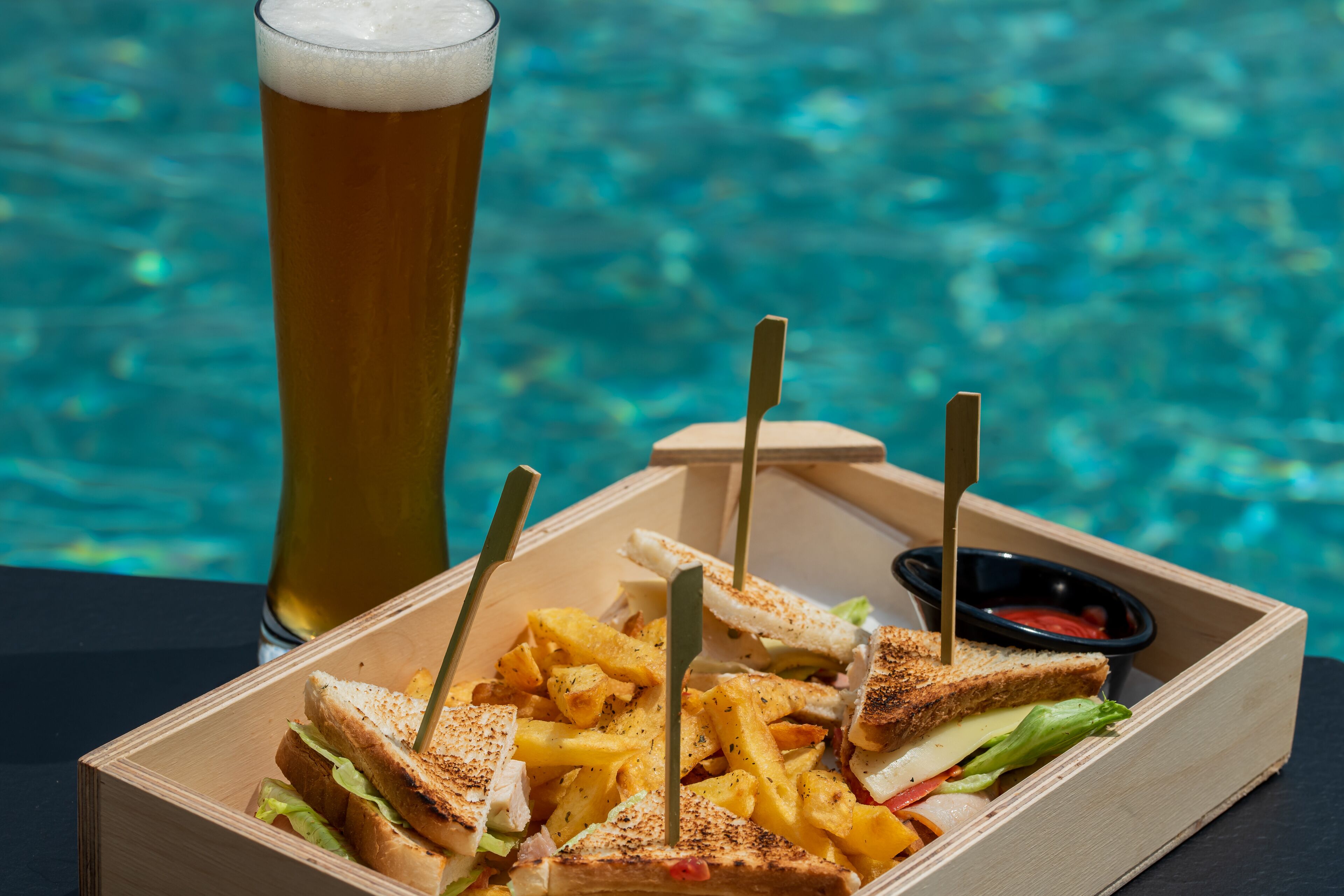 Un plateau repas avec des sandwichs au fromage grillé, des frites et un verre de bière, le tout servi au bord d’une piscine.