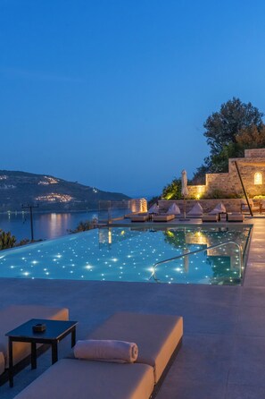 Un espace aménagé au bord de la piscine, avec des chaises longues, une table et une vue sur une étendue d’eau et des lumières lointaines.