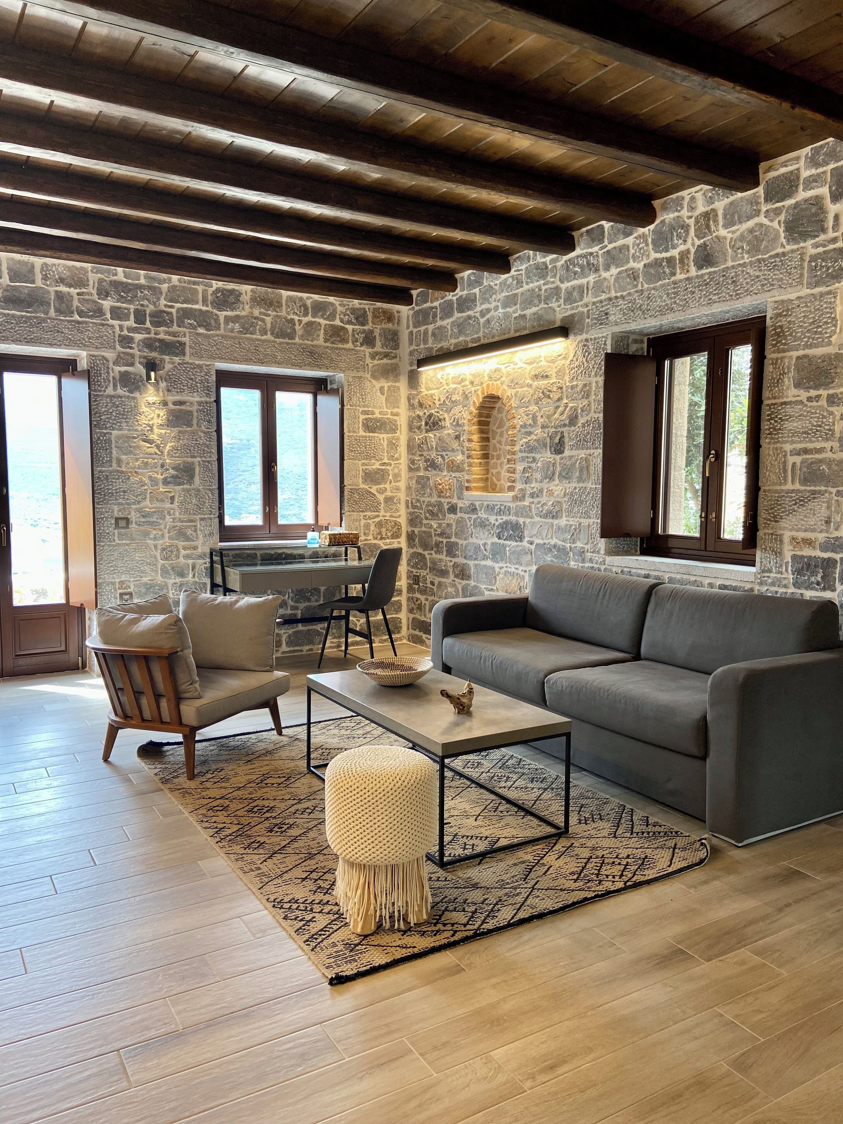 A cozy living room with a stone wall, a wooden ceiling, a grey sofa, a wooden chair, a coffee table, and a patterned rug.