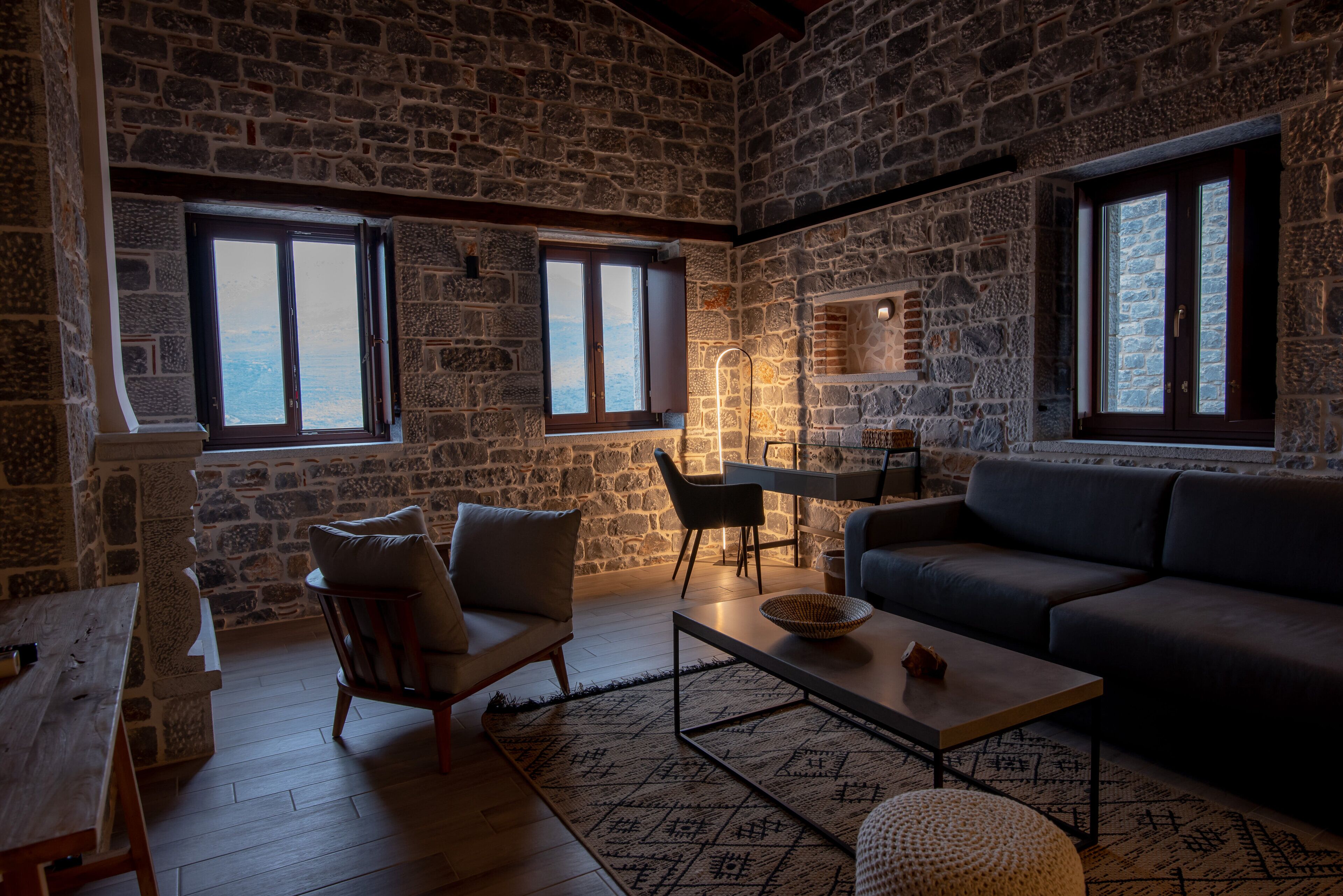 A stone-walled room with a sofa, chairs, a table, and a view through a window.