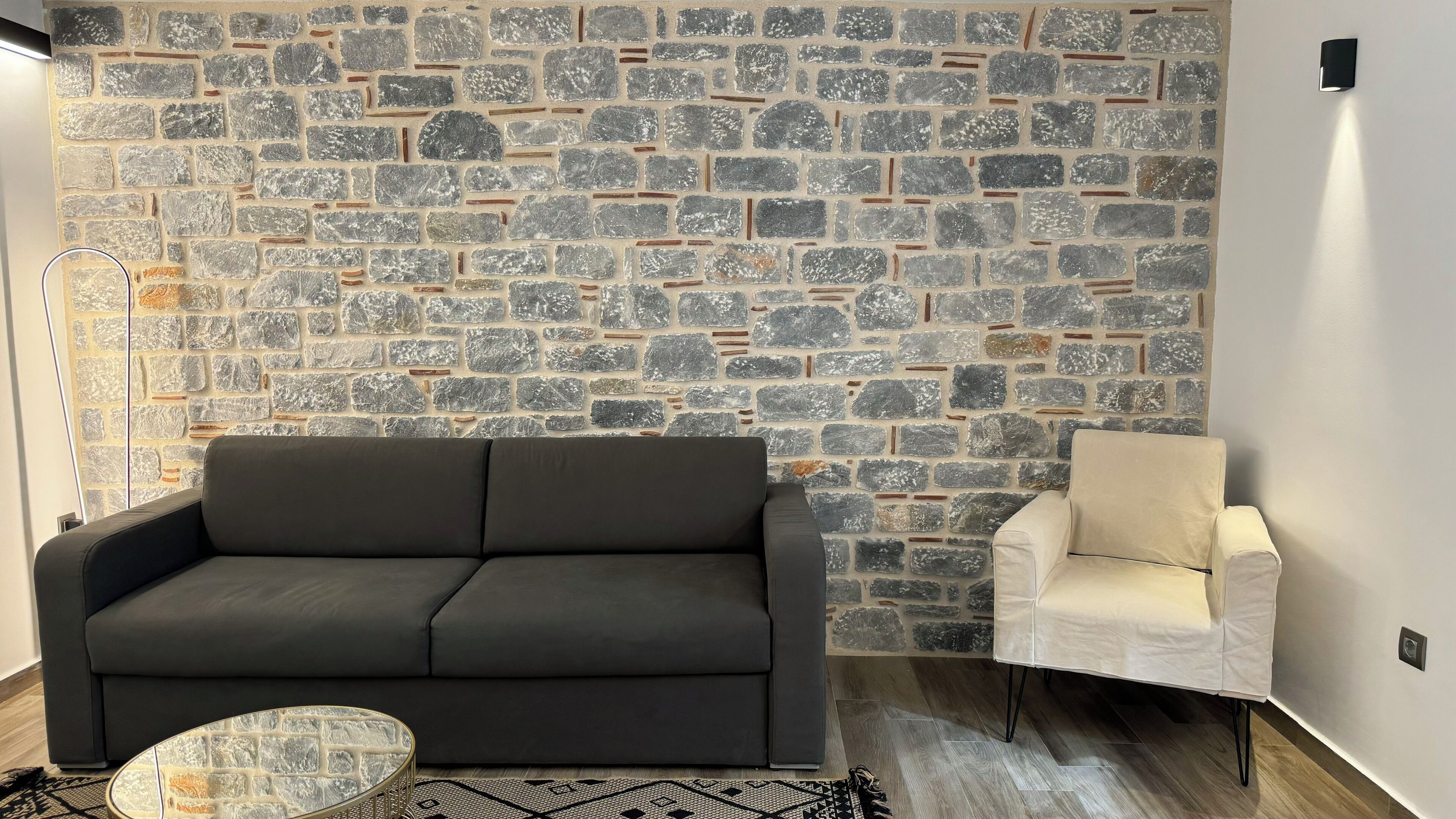 Une pièce avec un mur en pierre, un canapé sombre, un fauteuil blanc et une table basse ronde.