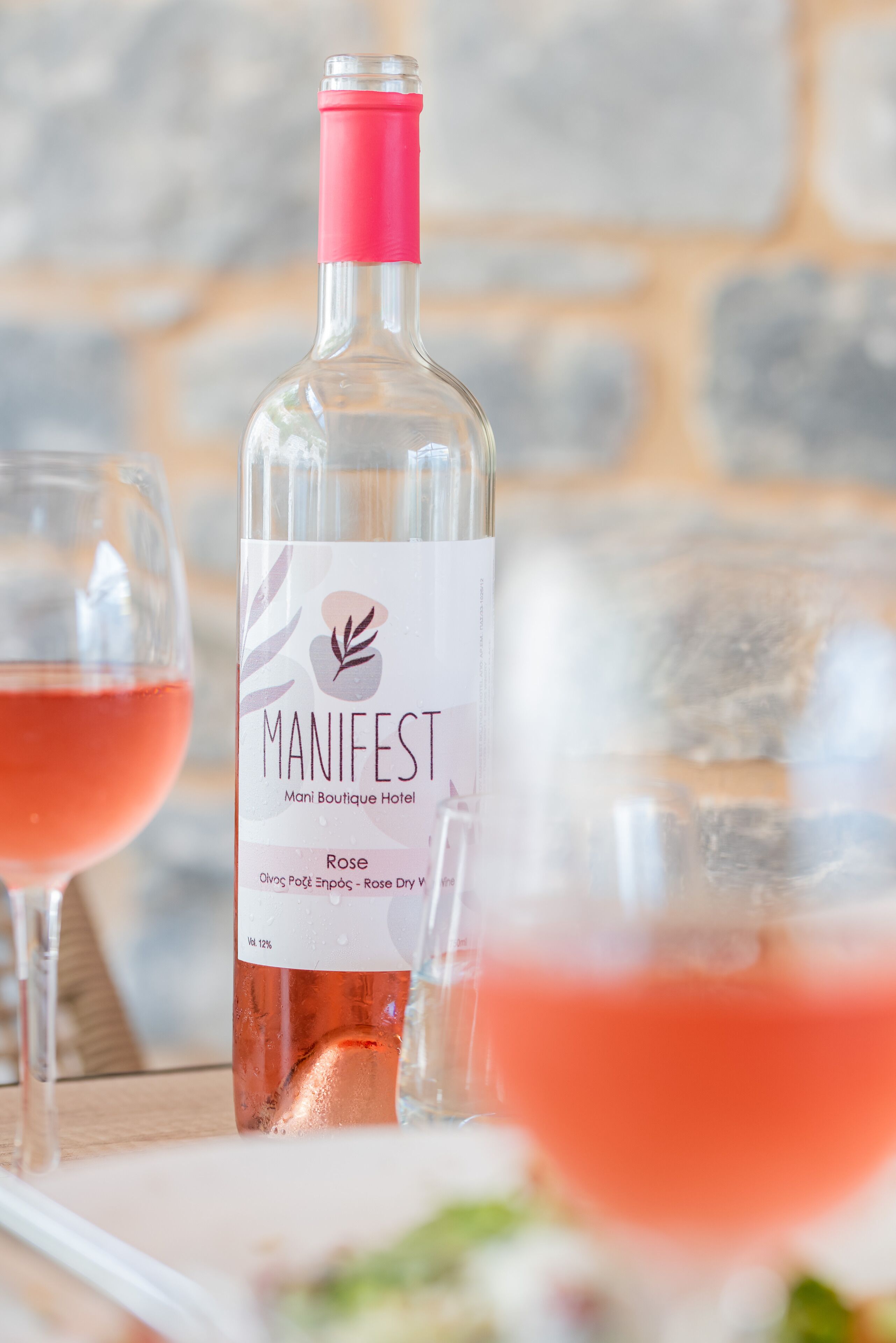 Une bouteille de vin Manifest avec une étiquette rose, deux verres de vin rosé et une table en bois.