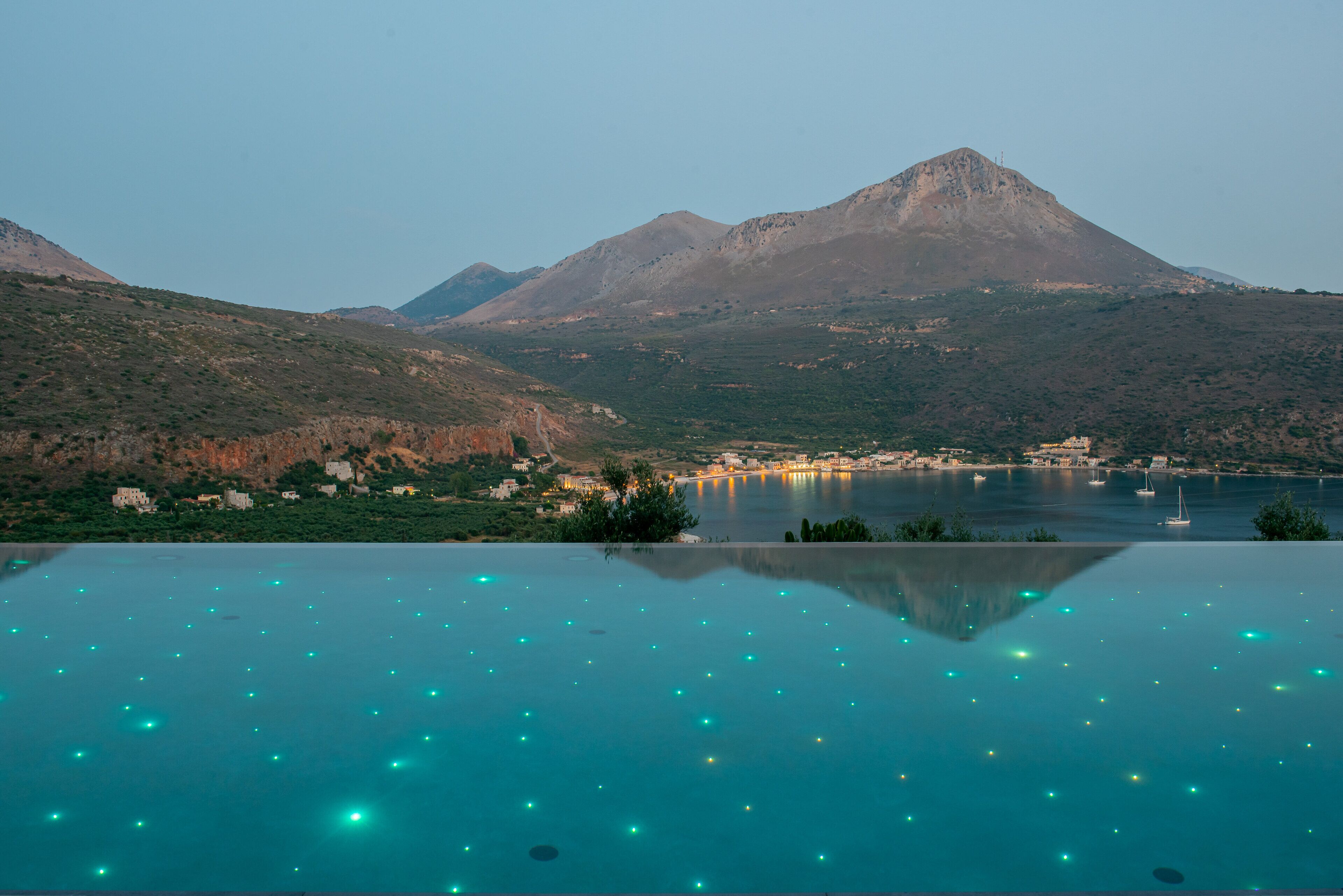 Vista panorámica de un paisaje montañoso con cielo despejado, un cuerpo de agua y una piscina con luces flotantes.