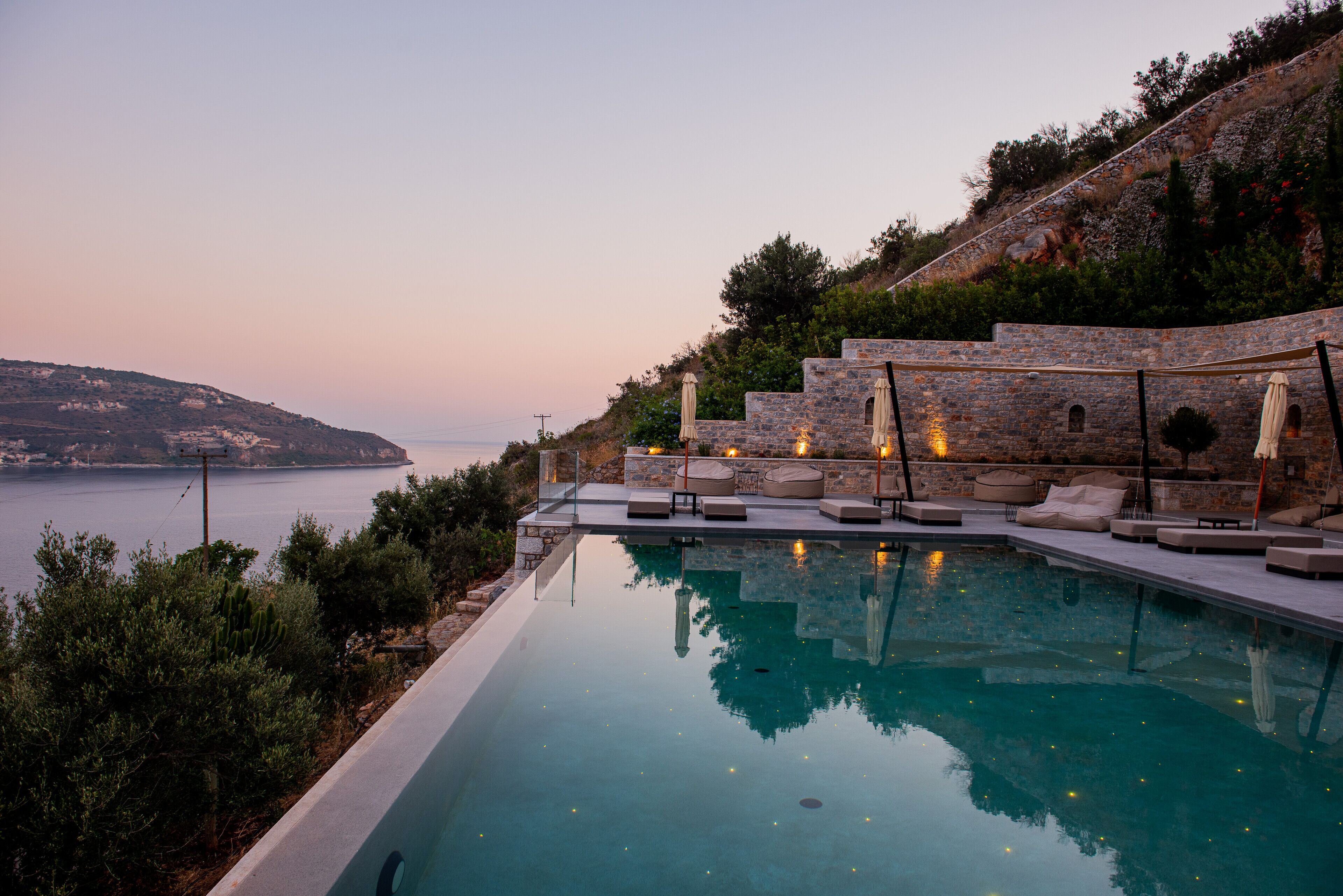 A poolside area with lounge chairs and umbrellas, overlooking a body of water and a hillside.
