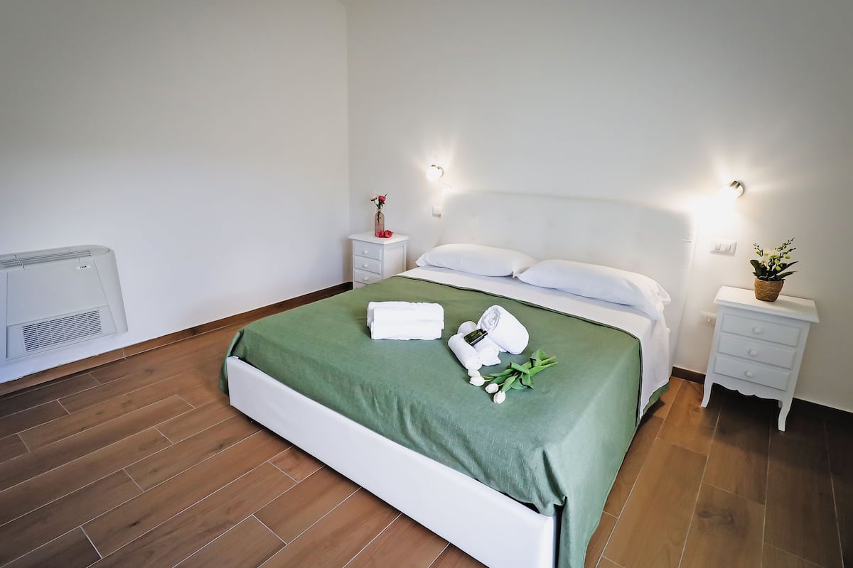 Ein ordentlich bezogenes Bett mit einem grünen Überwurf, weißen Kissen und einem weißen Nachttisch mit Blumenvase.