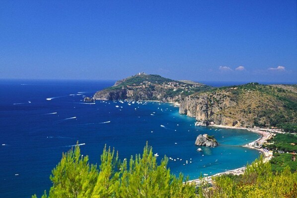 A coastal landscape with a rocky hill, clear blue sea, and boats.