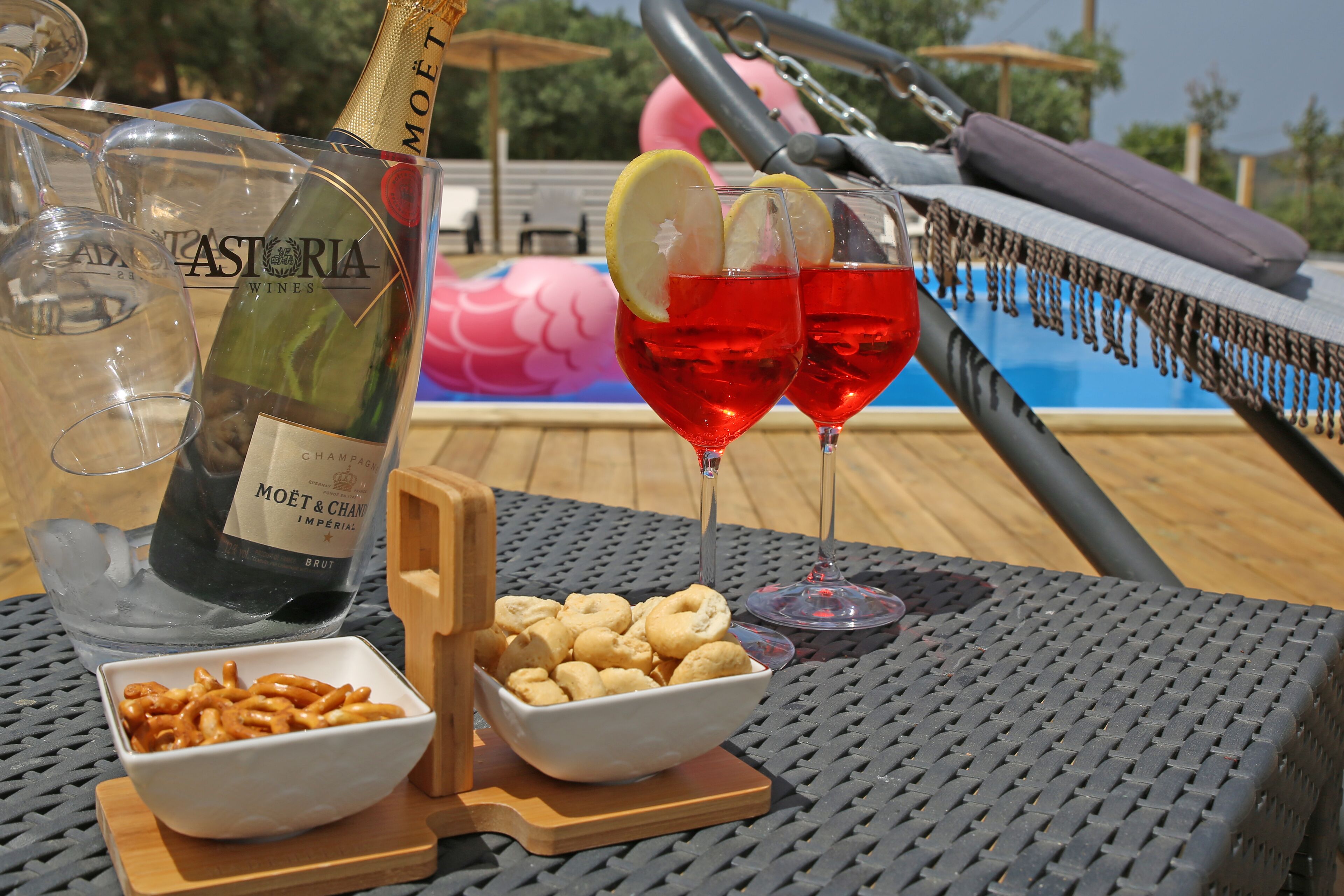 Ein Tisch mit einer Flasche Moët & Chandon Champagner, zwei Gläsern Rotwein mit Zitronenscheiben, einer Schale mit Snacks und einer Poolanlage im Hintergrund.