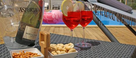 Ein Tisch mit einer Flasche Moët & Chandon Champagner, zwei Gläsern Rotwein mit Zitronenscheiben, einer Schale mit Snacks und einer Poolanlage im Hintergrund.