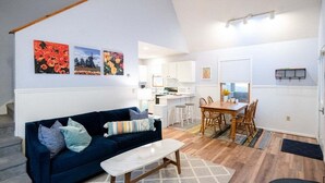 A modern living room with a dark blue sofa, a white coffee table, and a dining area with a wooden table and chairs. There is a kitchen in the background with white cabinets and a small island. - Goshorn Lake Resort Cottage #17 (Saugatuck)