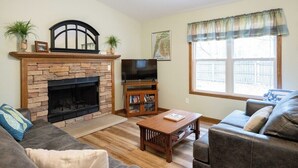 A living room with a stone fireplace, a television, a bookshelf, and a large window with a view. - Goshorn Lake Resort Cottage #2 (Saugatuck)