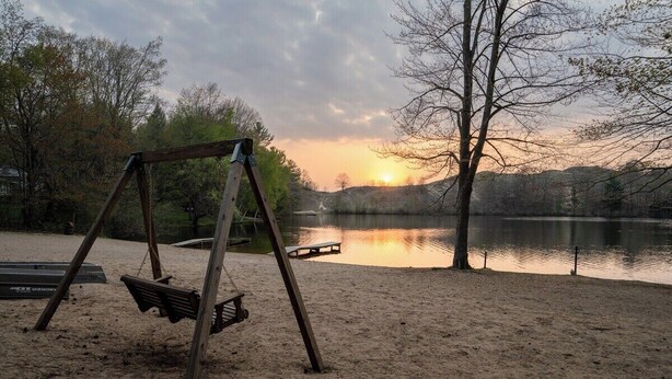Een houten schommelset op een zandstrand, aan een rustig meer, met een zonsondergang op de achtergrond.