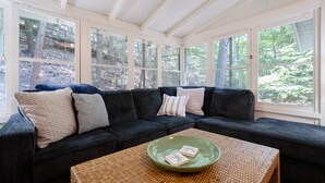 Una habitación con un sofá seccional de color oscuro, una mesa de centro tejida y ventanas amplias con vista a un bosque.