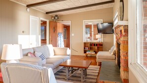 A living room with a sofa, armchair, coffee table, and fireplace. There is a television mounted on the wall and a bookshelf with books. - Lakefront Cottage | Private Beach + Sunset Views (Holland)