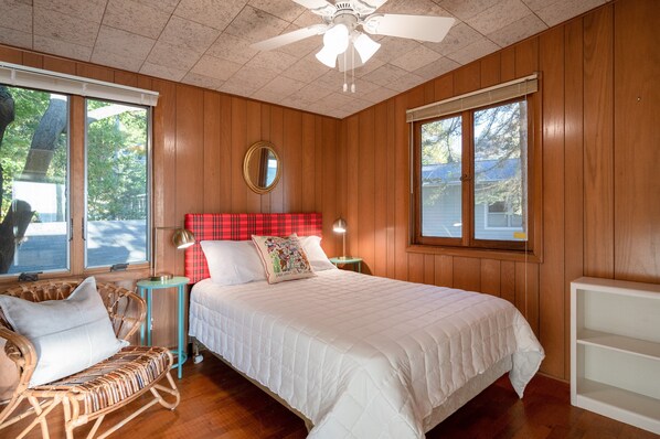 A bedroom with a wooden paneled wall, a bed with white bedding, a red plaid headboard, a wicker chair, and a small table with a lamp. - Lakefront Cottage | Private Beach + Sunset Views (Holland)
