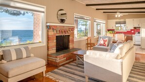 A cozy living room with a fireplace, large windows overlooking the sea, and a view into the kitchen. - Lakefront Cottage | Private Beach + Sunset Views (Holland)