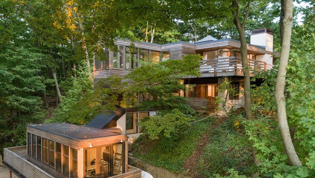 Une maison moderne dotée d’une terrasse fermée par des baies vitrées et d’une terrasse en bois, le tout entouré d’une forêt dense.