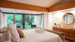 Une chambre avec un grand lit, une commode en bois, un miroir rond et une vue sur la verdure.