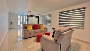 A spacious living room with a beige sofa, red ottoman, and a large window with blinds. - Luxury Villa with Pool, Heated Jacuzzi, & Terrace in El Yunque Rainforest (Río Grande)